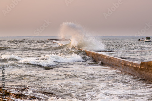 Fotografi Tempête marine, vague déferlante sur le jetée d'un petit port à la lumière rose d'un soir