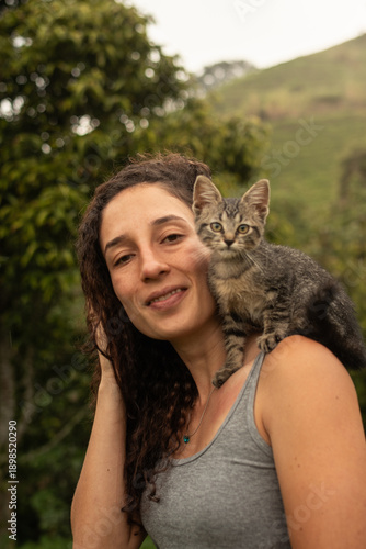 Cheerful young Hispanic woman looking directly at the camera with a soft smile while carrying a small tabby kitten on her shoulder. Rural nature background in Colombia.