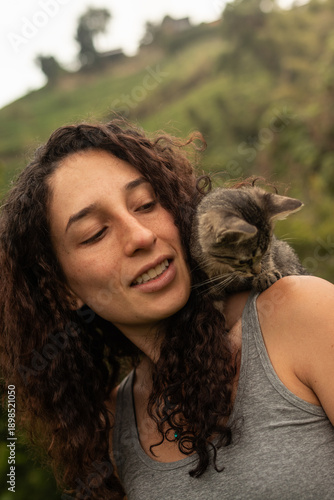 Cheerful Hispanic woman with eyes closed, enjoying a moment with a small tabby kitten interacting and pawing at her shoulder in a rural outdoor setting.