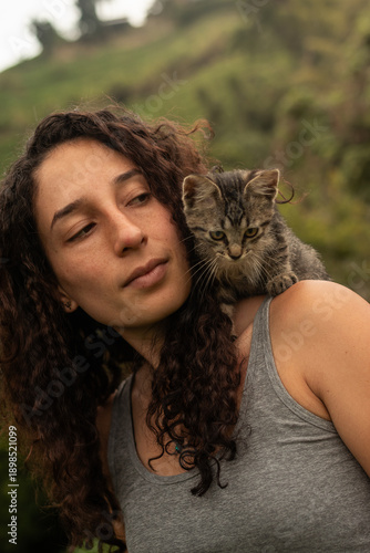 Portrait of a serious or contemplative Hispanic woman standing outdoors with a small tabby kitten on her shoulder. The kitten looks downwards while the woman looks at the horizon.