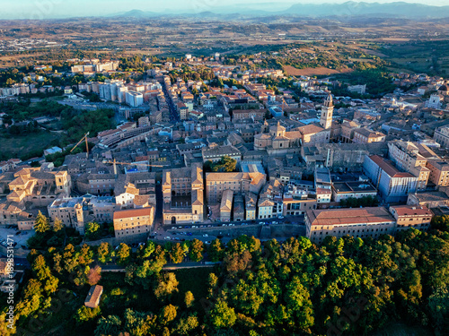 Aerial view of terracotta rooftops and sun-kissed facades meet verdant parks and ancient spires under a vast blue sky, Macerata, Italy.