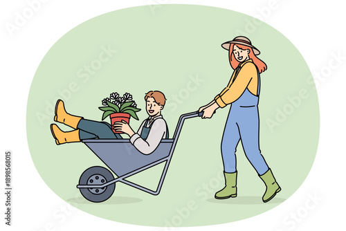 Woman farmer pushes man inside cart, enjoying good atmosphere in agro company team. Flowers in hands of farmer for planting seeds in fields and floriculture and getting large harvest