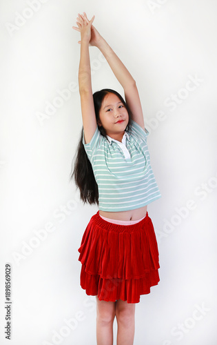 Portrait cheerful of young Asian girl in striped green shirt and a vibrant red tiered skirt standing on white background, gracefully raising her arms high in stretching pose.