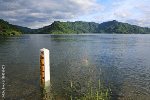 A water level gauge stands tall in the calm reservoir, monitoring the storage levels against a stunning backdrop of lush green mountains and a cloudy sky.