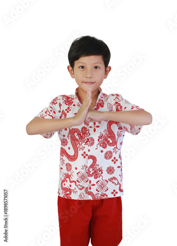 Cute young Asian boy wearing traditional white and red dragon pattern shirt, performing respectful Chinese greeting gesture (Gong Xi Fa Cai) on white background.