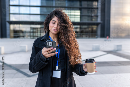 Woman with curly hair checking smartphone, drinking coffee, standing outside modern office building