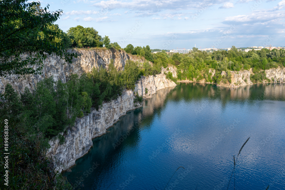 Fototapeta premium Wide landscape of zakrzowek quarry lake in Krakow, Poland, showing limestone cliffs, green trees, and calm blue water under a bright sky, representing urban nature escape, summer recreation, hiking vi