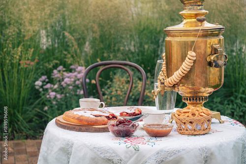 A golden samovar with bagels, tea cups, homemade pie, and jam on a white tablecloth set for tea in a garden.
