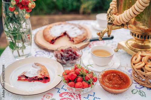 A rustic tea setting with golden samovar, fresh strawberries, jam, pie, and bagels arranged on an embroidered tablecloth outdoors.