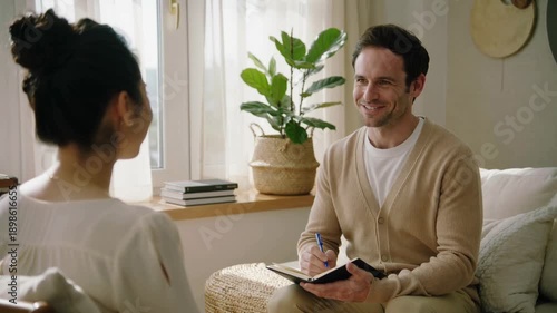 Male psychologist taking notes during counseling session with female client in bright and warm living room office.