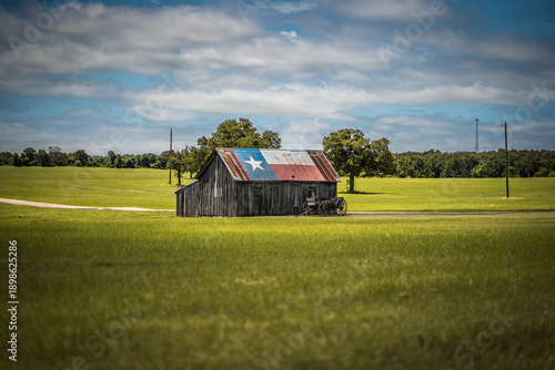 Old Rustic Barn With Texas Flag Painted on Roof With Old Wagon Nearby In Green Field in Rural Town in Texas