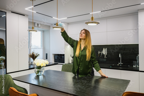 Woman wiping dust from a hanging lamp in a modern kitchen interior. Home cleaning routine, everyday household activity, minimalistic apartment, natural light.
