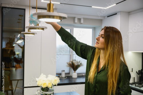 Woman wiping dust from a hanging lamp in a modern kitchen interior. Home cleaning routine, everyday household activity, minimalistic apartment, natural light.
