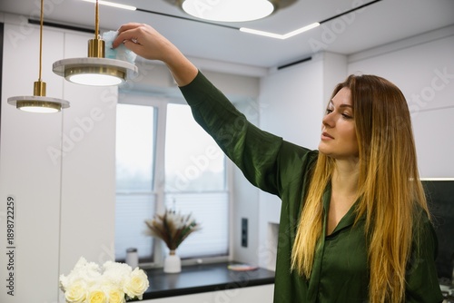 Woman wiping dust from a hanging lamp in a modern kitchen interior. Home cleaning routine, everyday household activity, minimalistic apartment, natural light.