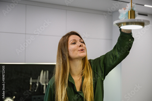 Woman wiping dust from a hanging lamp in a modern kitchen interior. Home cleaning routine, everyday household activity, minimalistic apartment, natural light.