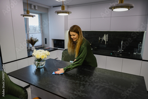 Adult woman cleaning dust from a kitchen island with a cloth in a modern home interior. Everyday housekeeping routine, minimalistic apartment, domestic cleaning activity.