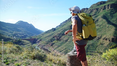 Man with a backpack looking at a panoramic mountain view and a dam in a valley, embodying healthy aging, exploration, and sustainable travel in nature