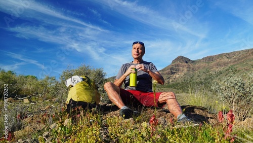 Active man in his 50s resting and drinking water from a bottle while hiking in a rugged mountain landscape under a clear blue sky, demonstrating a healthy and active lifestyle