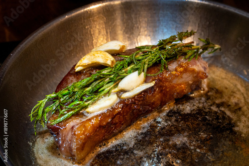 The chef cooks delicious meal on stove. The cook fries piece of juicy beef meat on frying pan with rosemary, pours oil using spoon. Process of steak cooking. Restaurant with professional kitchen.