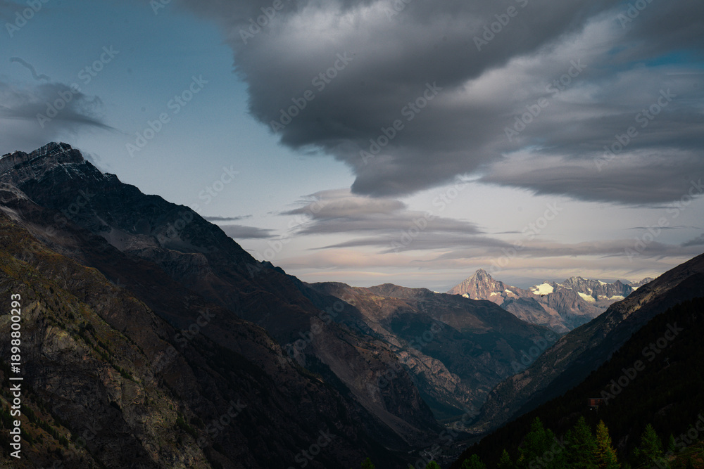 Obraz premium Dramatic mountain valley landscape under a moody cloudy sky in the Swiss Alps. Dark mountain slopes framing a distant sunlit peak at dusk. Cinematic alpine scenery near Zermatt, Switzerland.