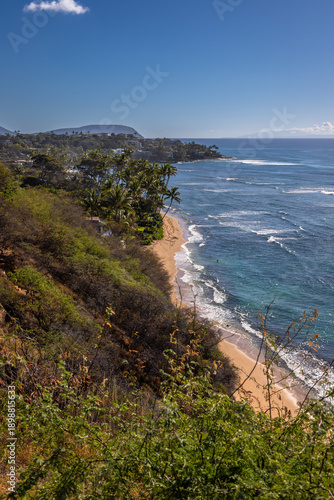 Scenic coastal view of Diamond Head Beach in Oahu, Hawaii, featuring turquoise ocean waves, sandy shoreline, tropical vegetation, and dramatic volcanic coastline under a clear blue sky.