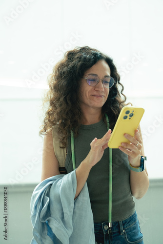 Close-up of smiling middle-aged curly brunette woman with glasses looking at smartphone, scrolling social media
