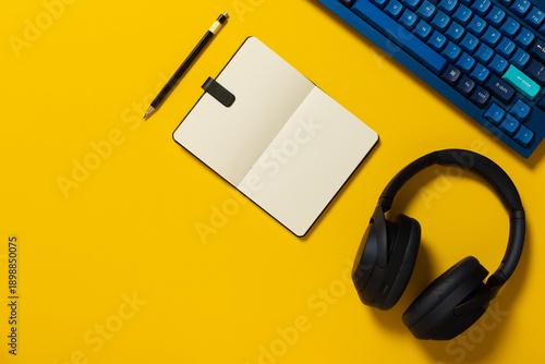 Overhead view of a mechanical keyboard, headphones, note pad and pen on colourful background