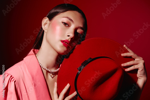 Fotografía Young woman with glossy red lipstick and blush poses holding a red hat in front of a red background