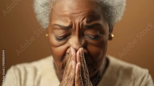 Middle-aged woman with white hair and wrinkles, hands clasped together in a prayerful pose, looking upwards. She seems to be focused on something or someone not visible in the video.