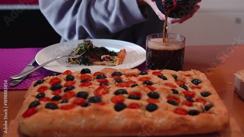 A young man pouring a drink into a glass to accompany his homemade Italian focaccia with black olives, cherry tomatoes, and rosemary