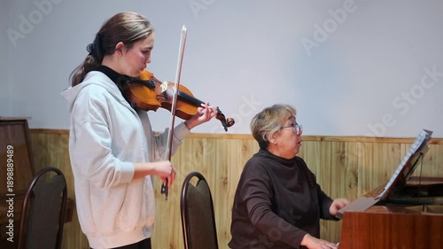 Teenage girl playing violin with piano accompaniment during a classical music lesson. Elderly teacher playing piano and watching the student attentively. Rehearsal in a music school classroom.