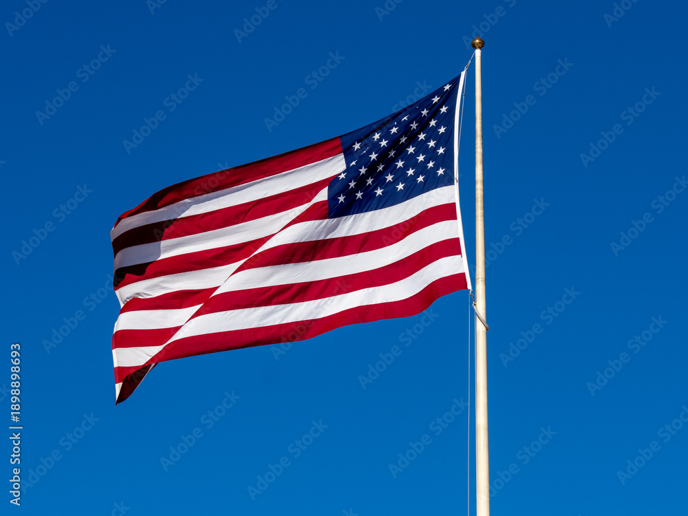 Fototapeta premium American Flag Waves Proudly in Clear Blue Sky Over a Tall Flagpole