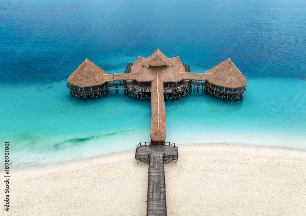 Naklejka premium Aerial top down view of a wooden pier with thatched roof over turquoise water in Zanzibar. Sea, bungalow, jetty on white sand beach in Nungwi, Tanzania. Luxury resort background for travel destination