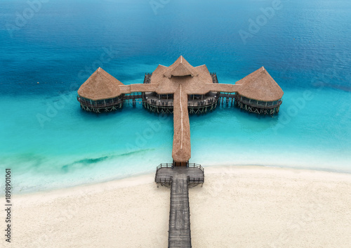 Aerial top down view of a wooden pier with thatched roof over turquoise water in Zanzibar. Sea, bungalow, jetty on white sand beach in Nungwi, Tanzania. Luxury resort background for travel destination