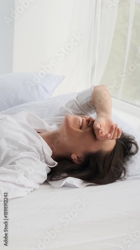 Vertical video of a happy young woman laughing while lying in a white bed in the morning. She is relaxed and enjoying a lazy start to the day.