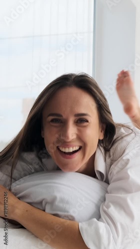 Vertical video of a happy young woman in a white shirt stretching and playfully falling onto a pillow in a bright bedroom, smiling at the camera.