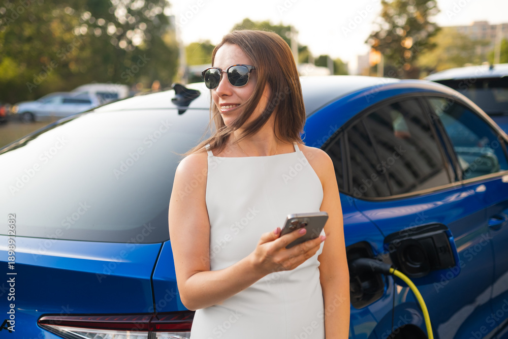 Naklejka premium Woman in sunglasses stands next to her electric car, checking her phone as it charges