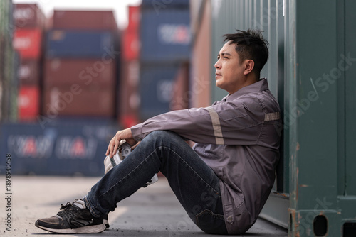A tired logistics worker in a gray uniform sits on the ground at a busy shipping container yard, holding his safety helmet while resting after a long shift in the freight transportation industry.