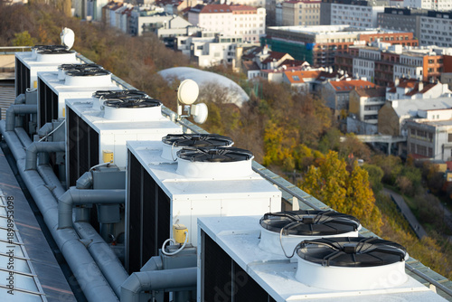 Rooftop view of multiple air conditioning units with fans and pipes, overlooking a cityscape with buildings and trees in the background during daylight