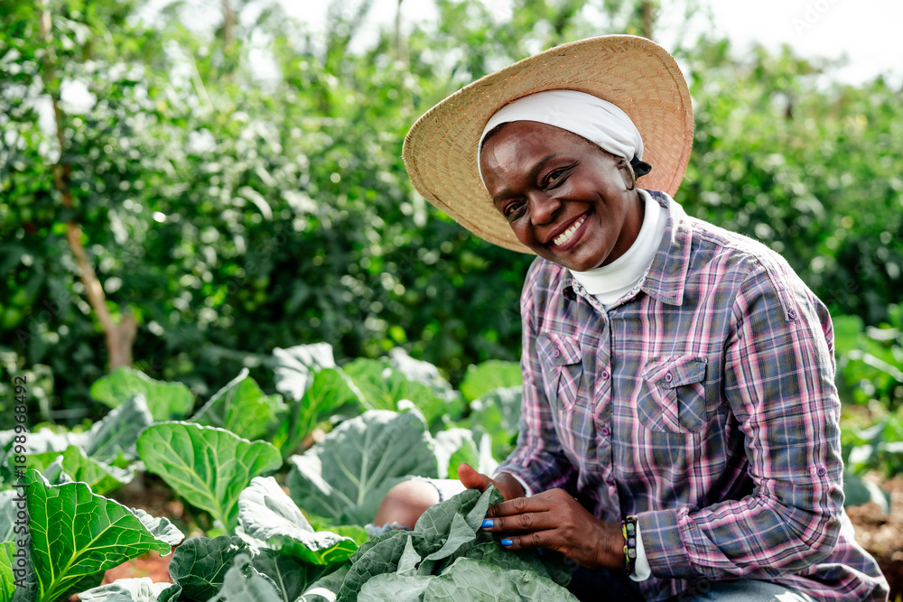Fototapeta premium Smiling senior African woman farmer working in an organic vegetable garden, wearing a straw hat and plaid shirt.
