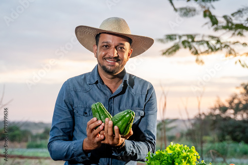 Fotografi Happy male farmer in a straw hat holding fresh chayote vegetables in a rural fie