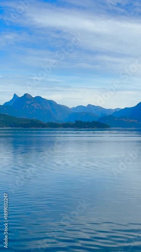 Vertical shot of a coastal scene taken from a passenger ferry on water, through rugged west coast of British Columbia, Canada, Mountain peaks and forest covered hillside with light streaks of clouds. 