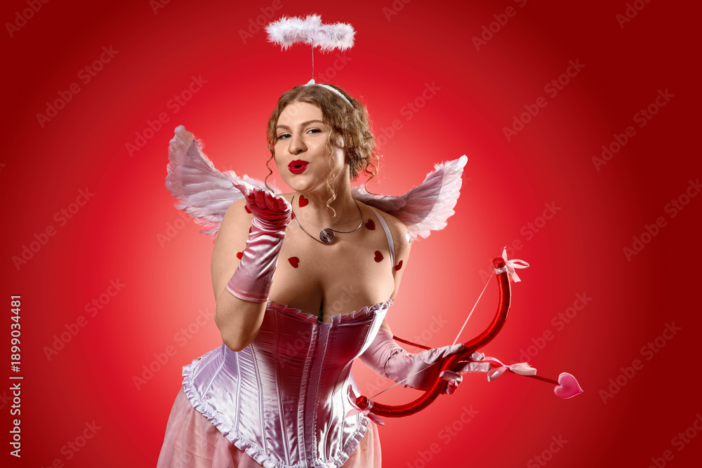 Fototapeta premium Beautiful woman dressed as Cupid with bow blowing kiss on red background. Valentine's Day celebration
