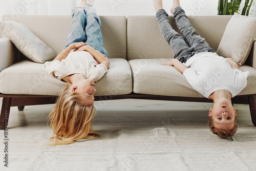 Two playful children lying upside down on sofa in casual clothes, enjoying relaxed moment at home with bright interior background.