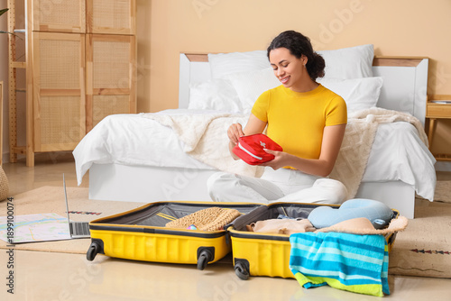 Bild auf Leinwand Young African-American woman packing first aid kit into suitcase in bedroom