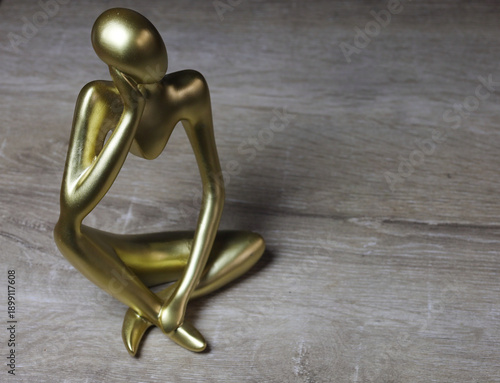 Golden Abstract Sculpture of Contemplative Human Figure. A modern golden sculpture depicting a stylized seated human figure in a thoughtful pose. Crafted with smooth, elongated limbs and a minimalist 
