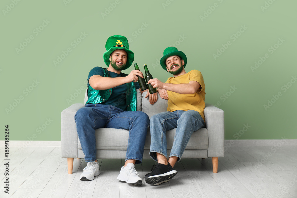 Naklejka premium Young men in leprechaun hats with green beards holding bottles of beer and sitting on sofa near green wall. St. Patrick's Day celebration