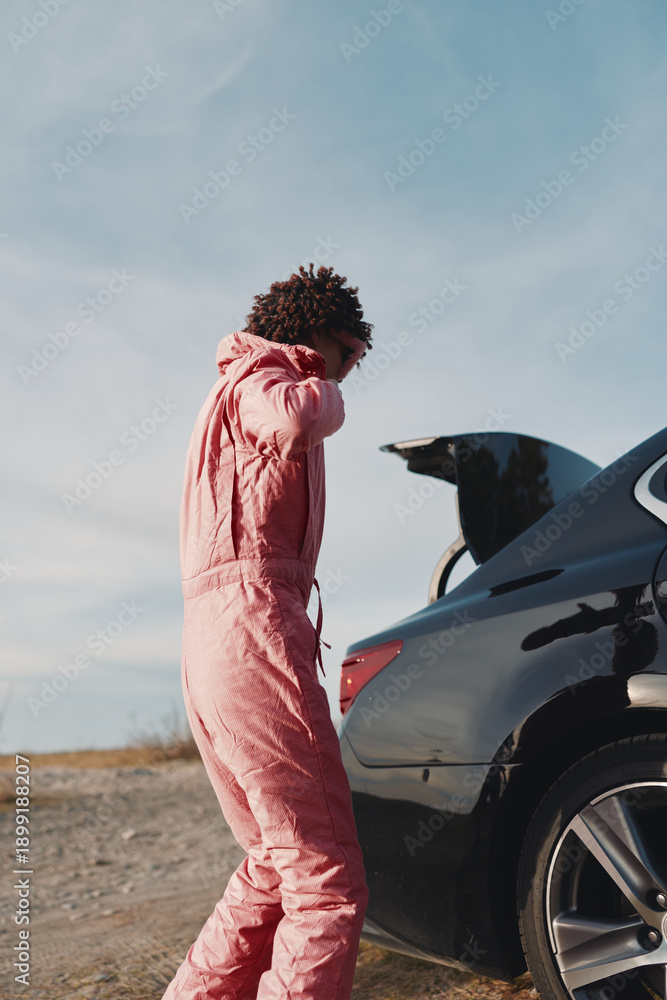 Obraz premium Car trunk open, outdoors scene with a person wearing a pink jumpsuit by a road in a desert, preparing belongings under a clear sky and bright sunlight.