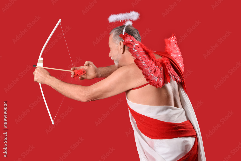 Fototapeta premium Mature man dressed as Cupid shooting bow on red background. Valentine's Day celebration