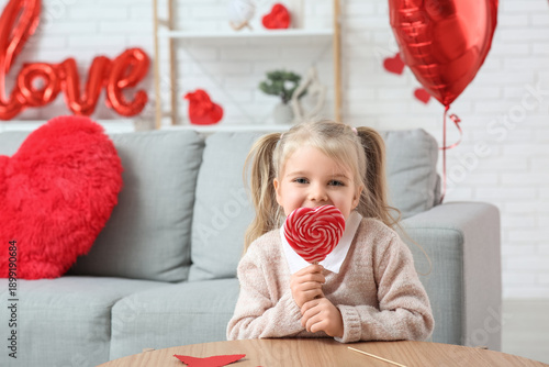 Cute little girl with lollipop sitting in room decorated for Valentines Day